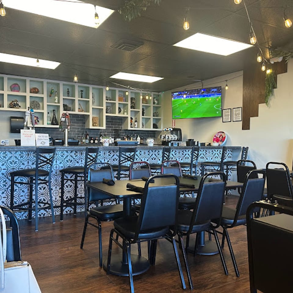 Interior of Itacate Mexican Food, showing a bar with patterned tiles, dining tables, and a TV playing soccer.