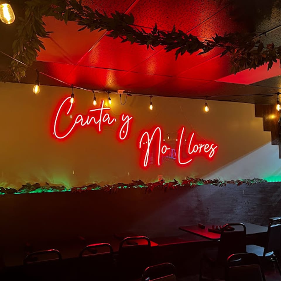 Red neon sign "Canta y No Llores" on a wall with string lights and green foliage.