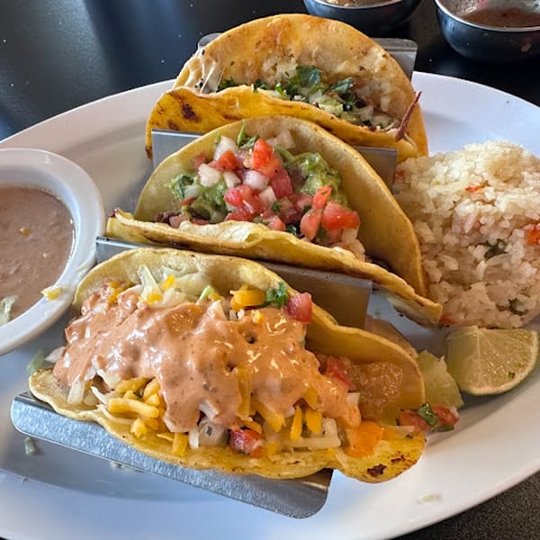 Three Itacate Mexican Food tacos with rice, beans, and lime wedges on a white plate.