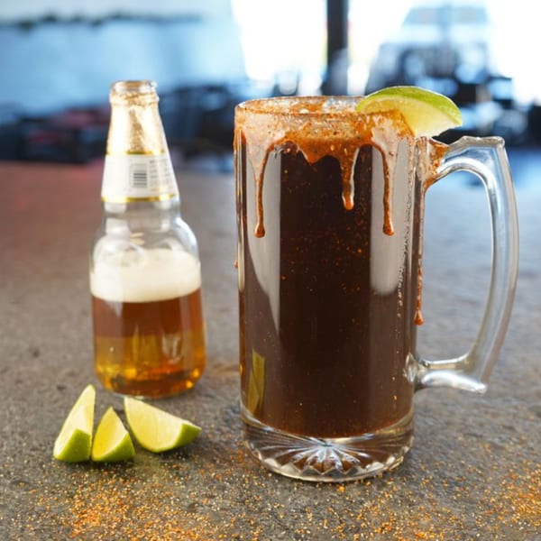 A dark michelada in a mug with a lime wedge, next to a beer bottle and lime slices.