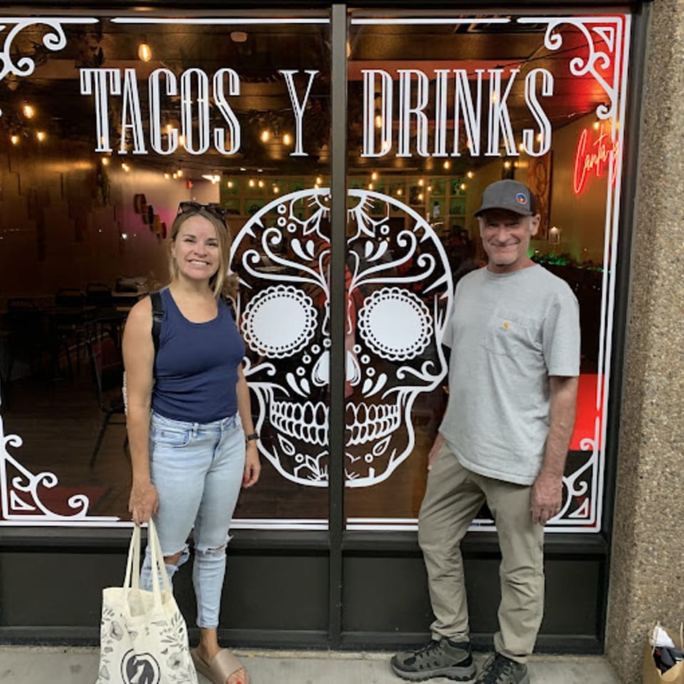 Two people stand outside Itacate Mexican Food, featuring a sugar skull and "Tacos y Drinks" on the window.